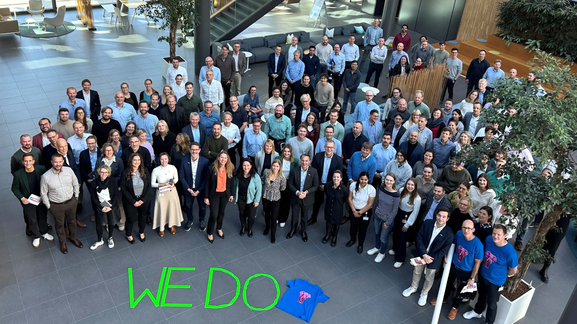 Sustainability Week 2025 A large group poses in a sunlit atrium. A blue t-shirt and the words "WE DO" in green, written with tesa tape, are in front. (This text has been generated by AI)