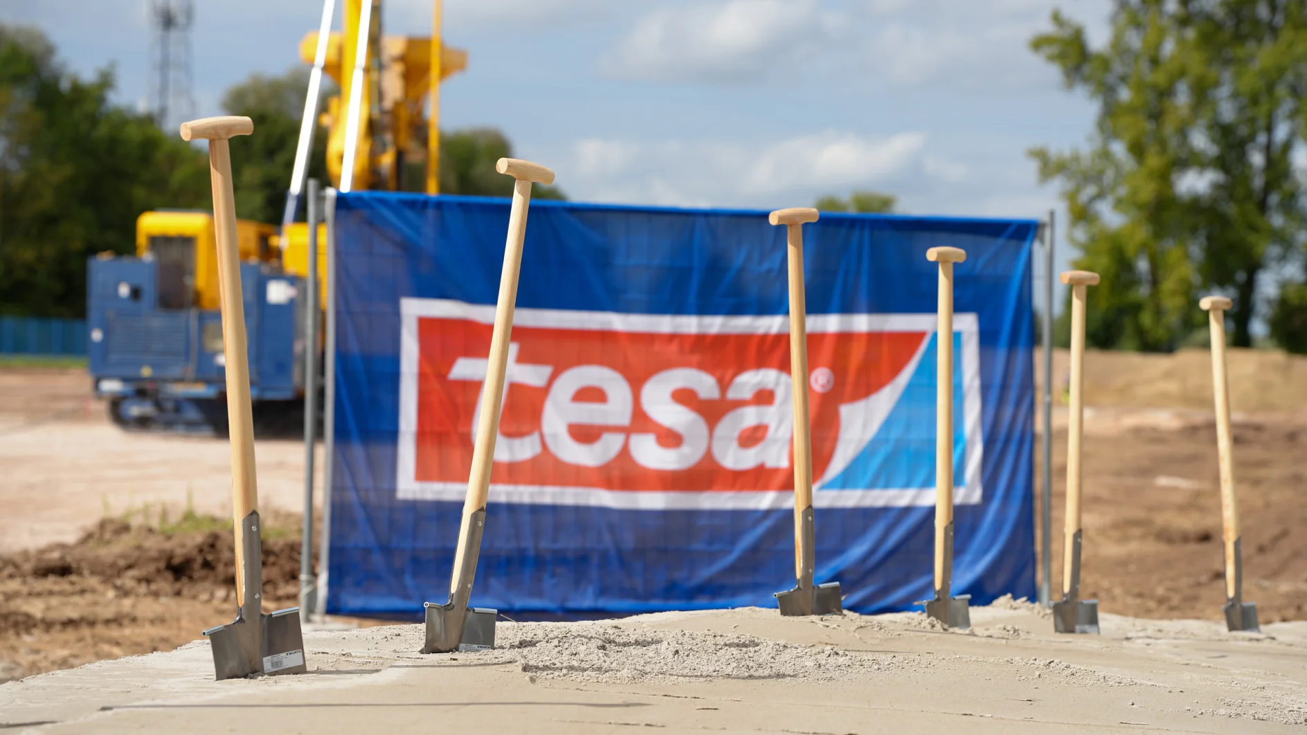 Construction start of the plant expansion for more capacity and future security: New production building at the tesa site in Offenburg is scheduled to go into operation in 2027. Six shovels stand in a concrete slab at a construction site. A blue tesa tape banner, equipment, trees, and clouds are visible. (This text has been generated by AI)