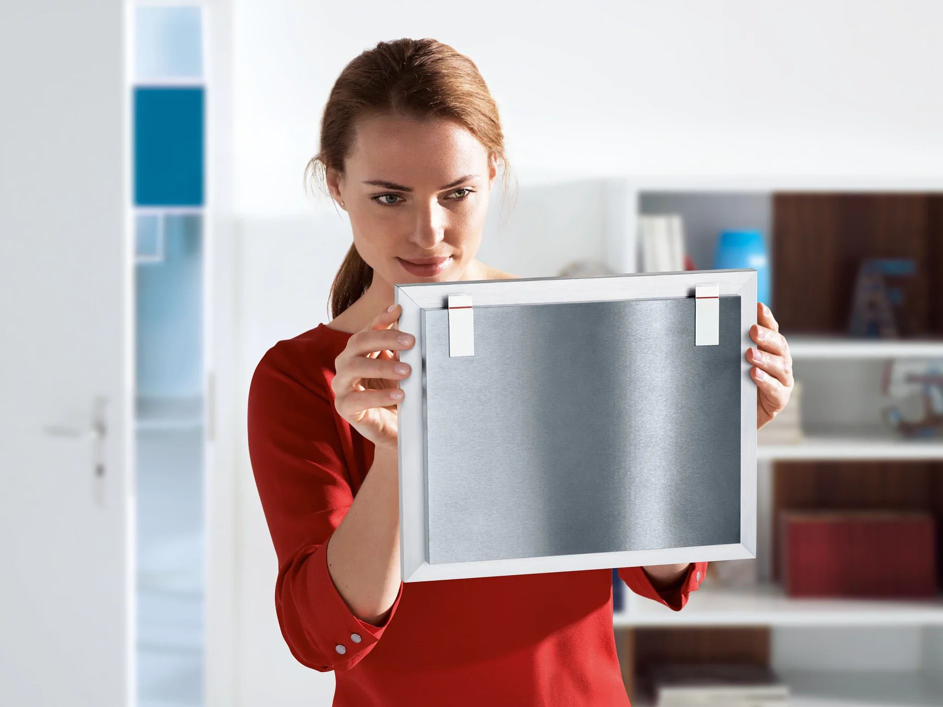 Stage (1) A woman in a red shirt examines a framed metal object, holding it in both hands. She stands in a brightly lit room with a bookshelf in the background and an open door to the left. The frame is secured with tesa tape, offering reliable and sturdy support for her display. (This text has been generated by AI)