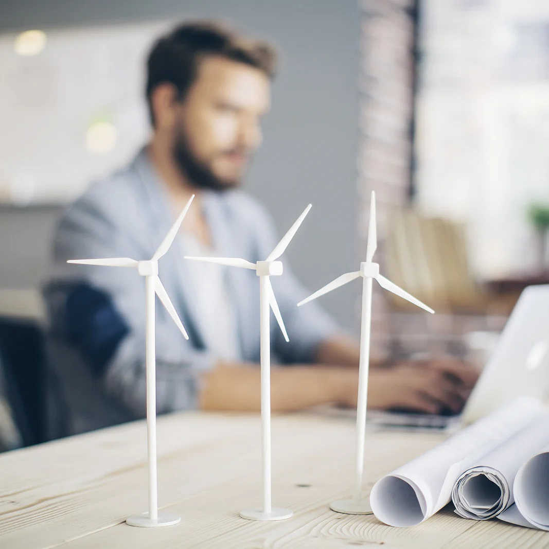 SIM_Wind-Energy-Screen Üç küçük rüzgar türbini modeli, rulo haline getirilmiş planların yanındaki bir masanın üzerindedir. Arka planda, dizüstü bilgisayarlı bir kişi bulanık ve odak dışı bir şekilde oturmaktadır, bu da yenilenebilir enerji tasarımı veya mühendisliğiyle ilgili bir çalışma alanı ortamını ima eder. (This text has been generated by AI)