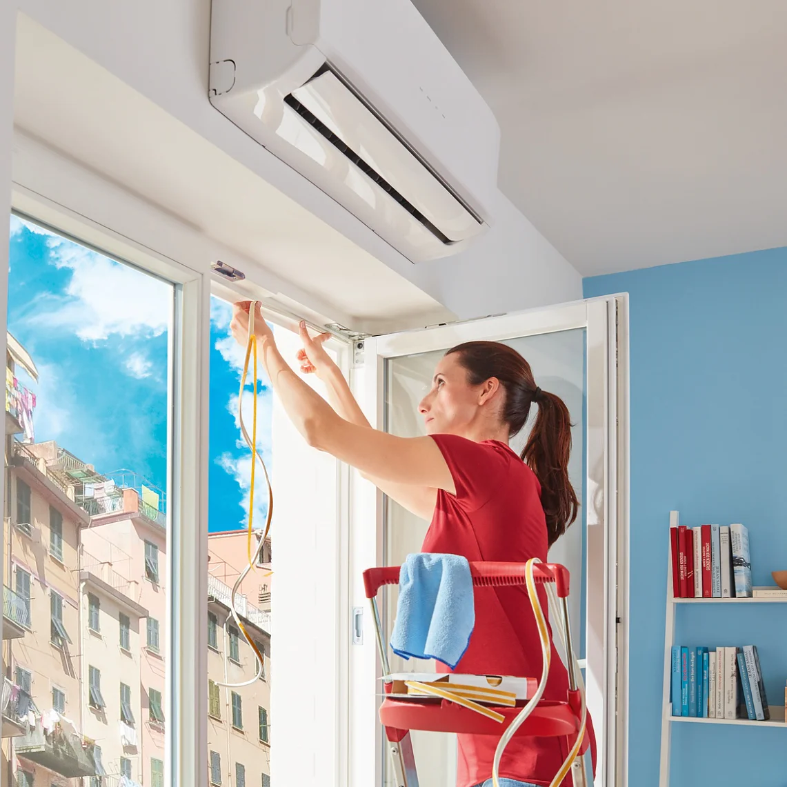 tesamoll-p-profle_kv_header_001-Screen Merdivendeki bir kadın bir kapının üstüne bir klima ünitesi takıyor. Uzaktan kumandayı tutarken kabloları ayarlıyor. Odanın rafları kitap ve dekoratif eşyalarla dolu mavi bir duvarı var. Güneş ışığı açık kapıdan içeri sızıyor ve bir şehir manzarası ortaya çıkıyor. Kabloları etkili bir şekilde yerinde tutmak için yakınlara yerleştirilmiş tesa bant ruloları var. (This text has been generated by AI)