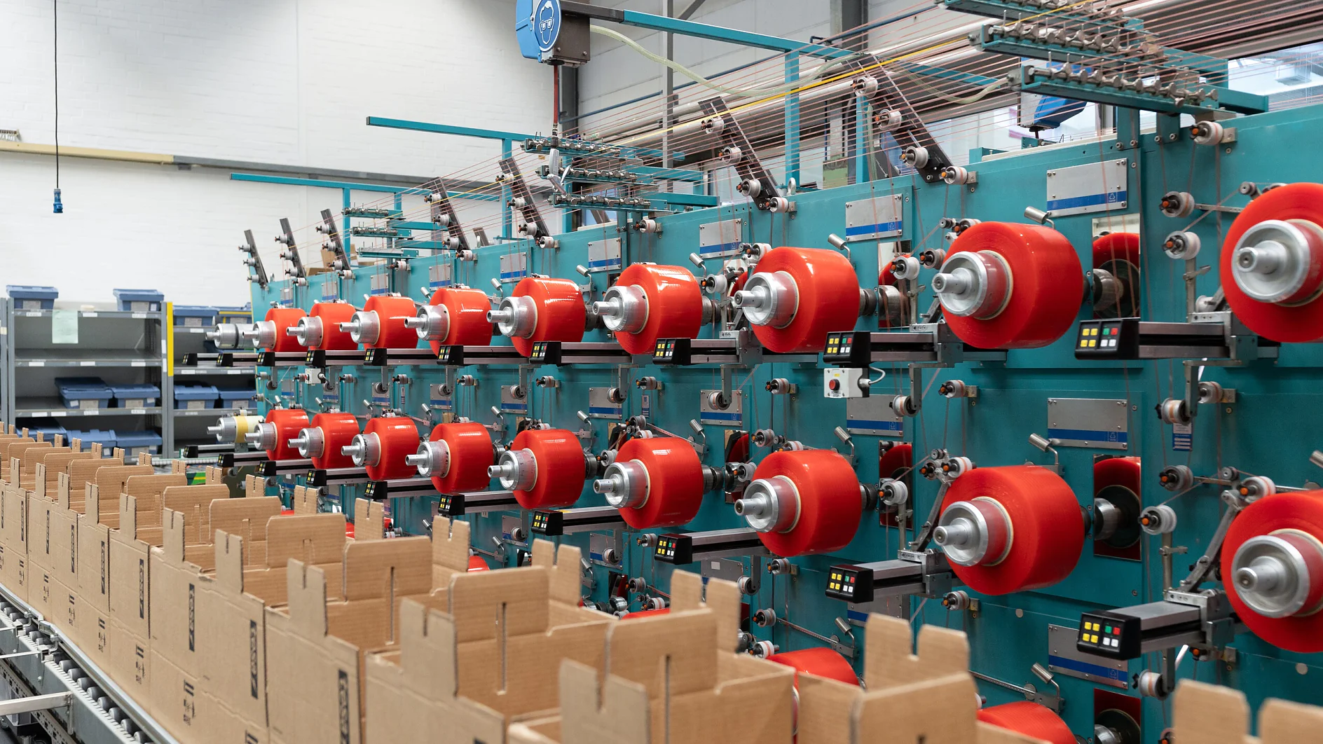 tWHH, bant üretimi, makine, bant, tesis, saha, Sanayi, üretim süreci, Mood, üretim, yapışkan bant_75-cms A factory interior showing a row of teal-colored machines with multiple red spools of thread and rolls of tesa tape. Cardboard boxes are aligned on a conveyor belt in the foreground. The setting appears organized, with equipment and materials neatly arranged. (This text has been generated by AI)