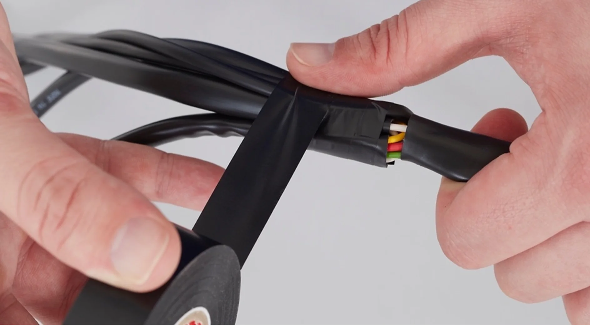 Temel Paketleme Bir kişi bir grup renkli kablonun etrafına siyah tesa bant sarıyor. Tesa bandı yeşil, sarı ve kırmızı renkleri içeren kabloları sabitlemek ve yalıtmak için uygulanıyor. Arka plan düz beyazdır. (This text has been generated by AI)