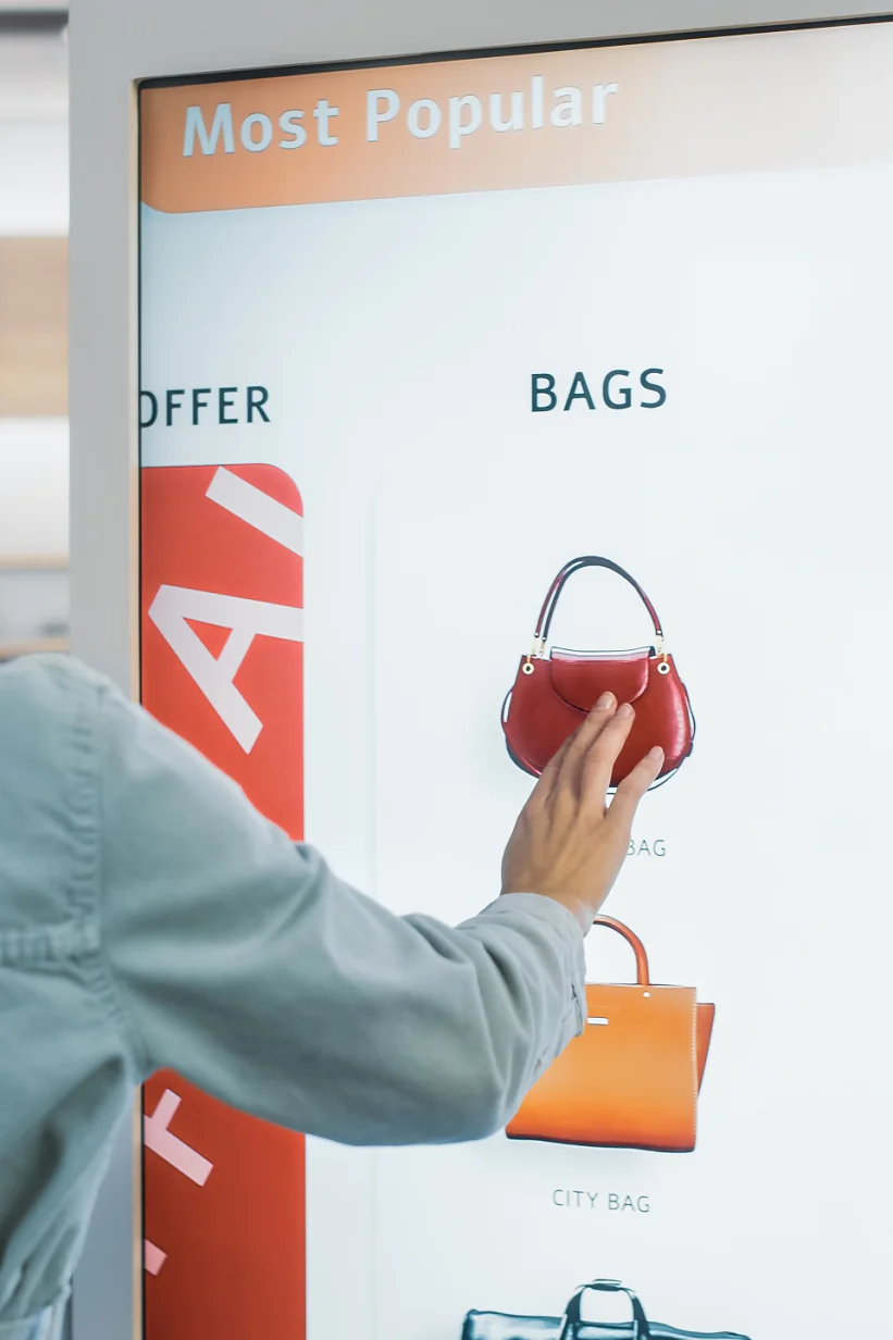 Smuk kvindelig kunde, der bruger gulvstående LCD-touchskærm, mens hun handler i tøjbutik. Hun udvælger stilfulde tasker og vælger forskellige designs fra kollektionen. Folk i moderigtig butik.