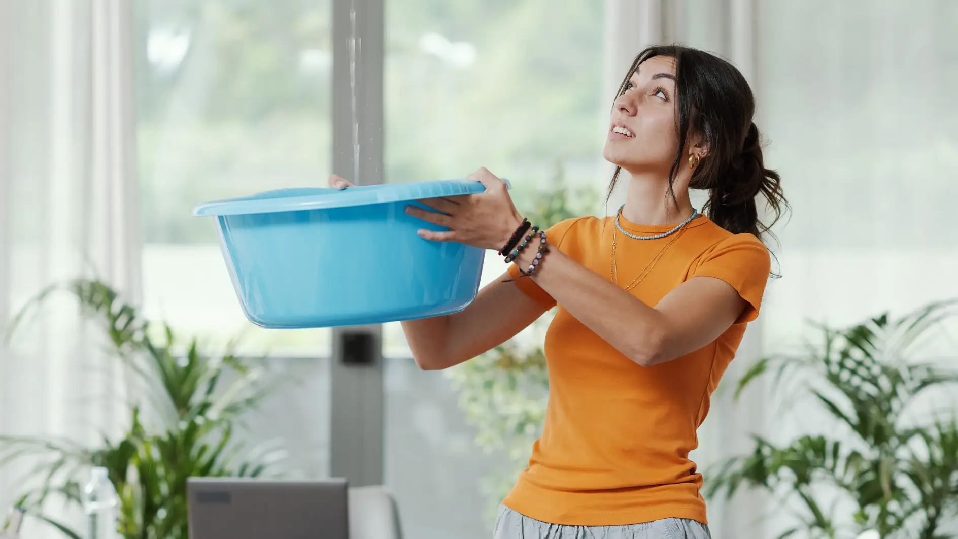 Young upset woman collecting water leaking and dripping from the ceiling