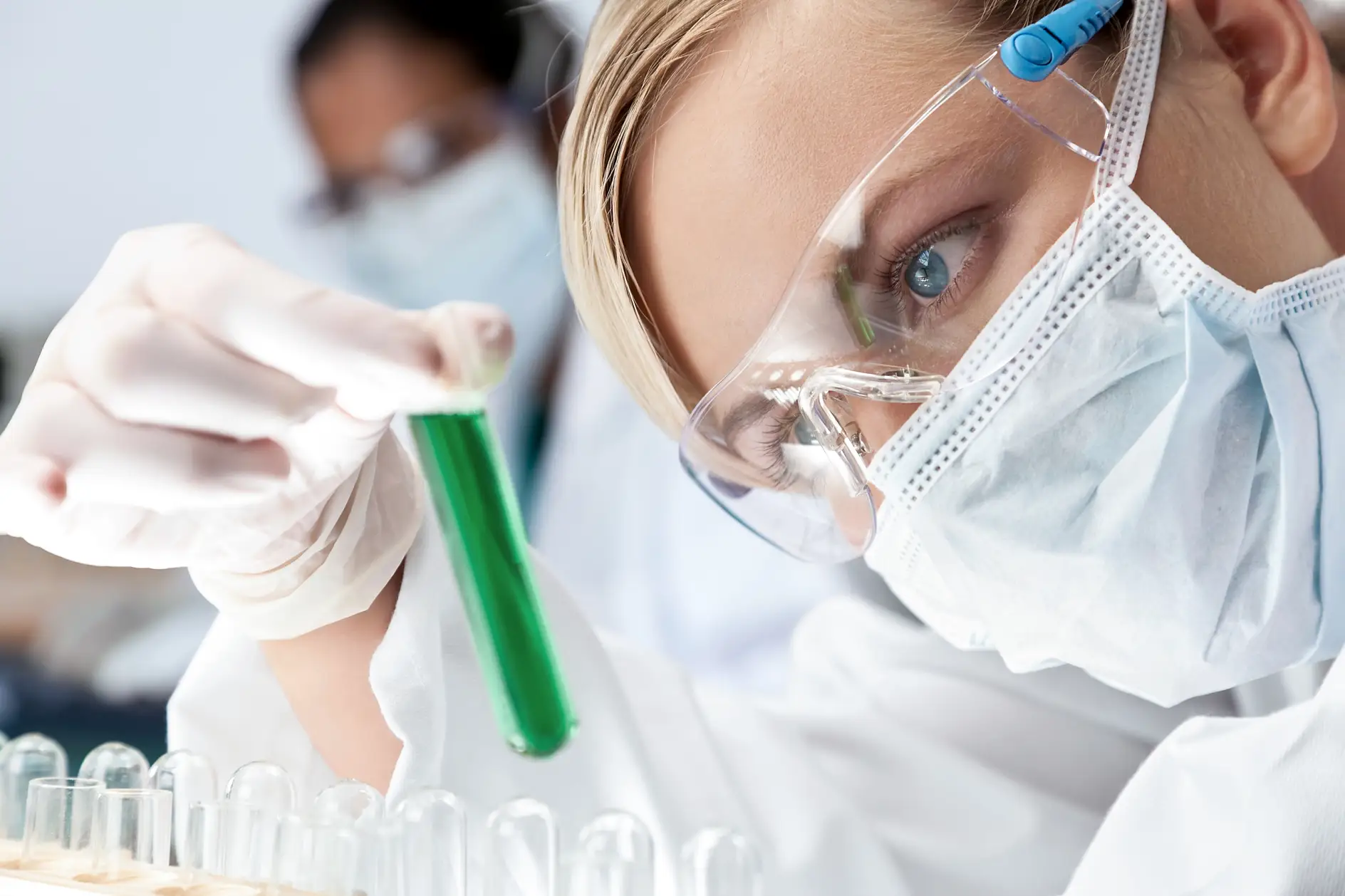 Une femme médecin ou chercheuse scientifique regardant une solution verte dans un laboratoire, avec sa collègue asiatique dans le flou derrière elle.