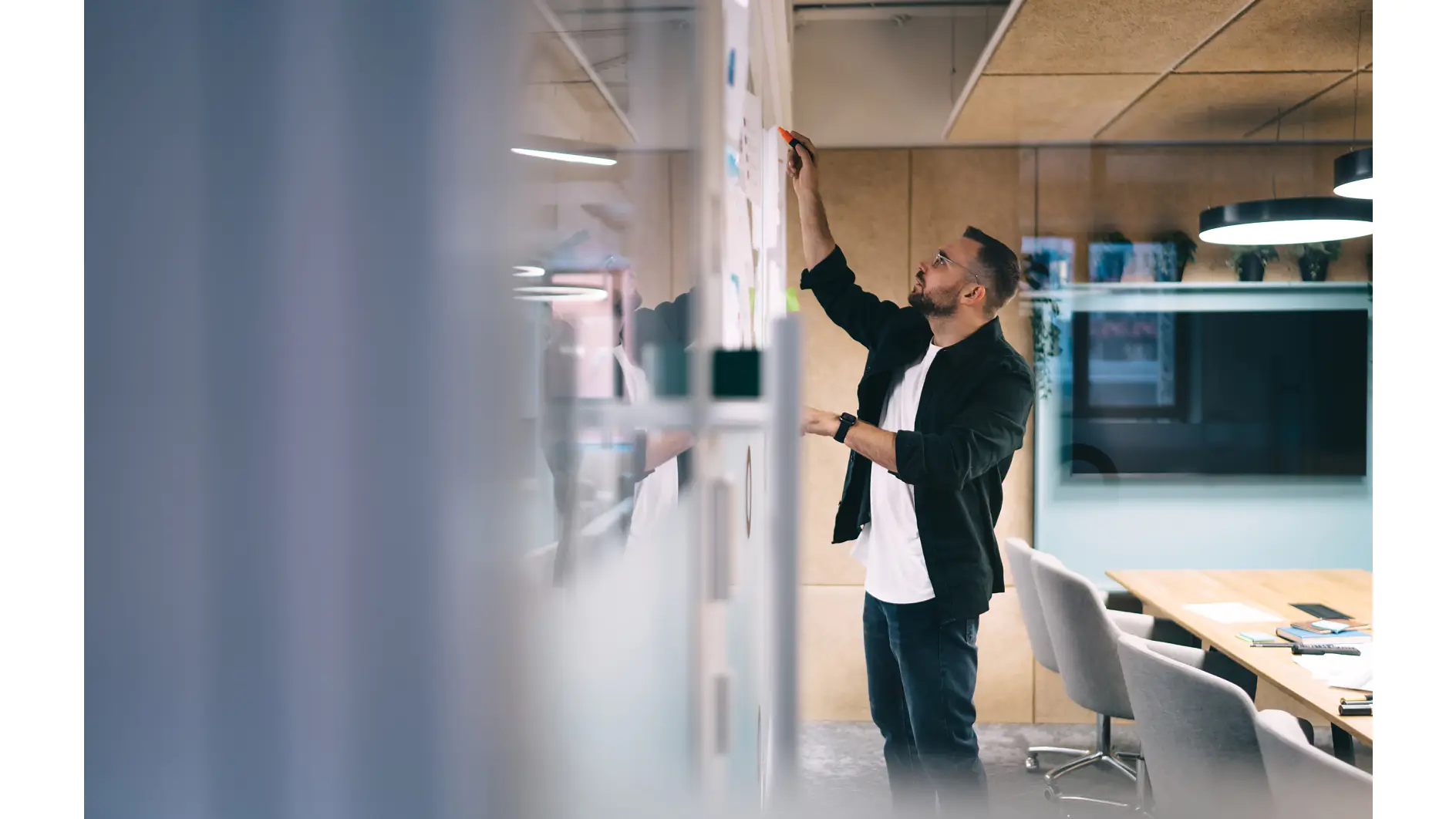 Side view of concentrated bearded male employee in casual clothes standing near glass in modern creative workspace and drawing scheme with marker