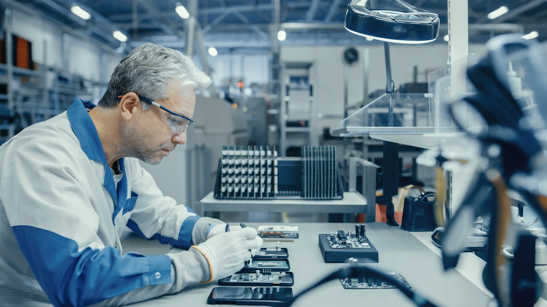 Senior Man in Blue - White Work Coat is Using Plier to Assemble Printed Circuit Board for Smartphone. Electronics Factory Workers in a High Tech Factory Facility.